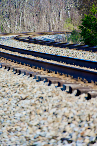 A set of curved railroad tracks slanted toward the inside of the curve atop a gravel railroad bed and railroad ties in a forested area on a sunny day.