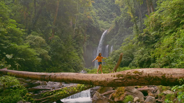 Woman crossing river by log on the background of tropical waterfall while hiking