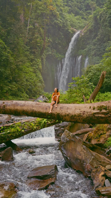 Woman sitting on huge log on the background of tropical waterfall while hiking