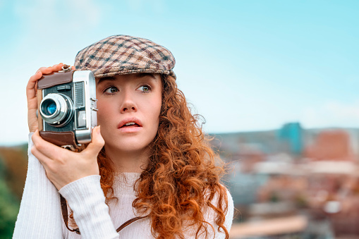 Portrait of an happy young redhead woman with red curly hair exploring a city and taking pictures with an old film camera.