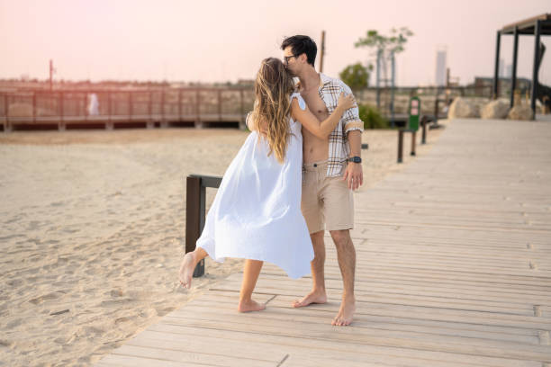 joven pareja enamorada abrazándose mientras está de pie en la pasarela de la playa al atardecer. hombre guapo besando a su novia. - honeymoon beach couple heterosexual couple fotografías e imágenes de stock