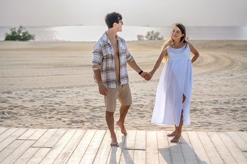 Young pregnant couple holding hands and walking on the wooden walkway relaxed and talking about future plans on the beach at sunset.