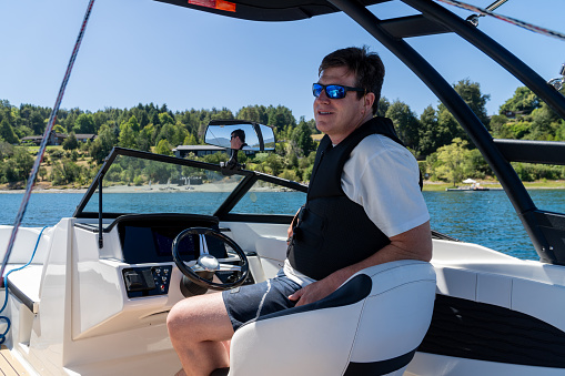Man wearing sunglasses, a life jacket, and swimwear, enjoying peace on a summer day at the lake while sitting on his boat.