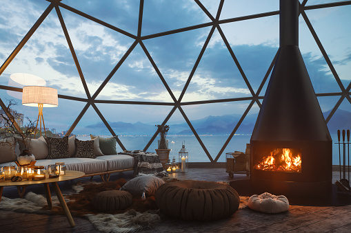 Interior of a geodesic dome house with a cozy fireplace and cushions. 
Panoramic windows offer views of the seascape and mountainous terrain, seamlessly blending the comforts of home with the beauty of nature.