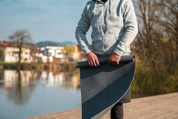 Close-up of a person ready for a workout, holding a yoga mat beside a calm lake with reflections and clear sky.