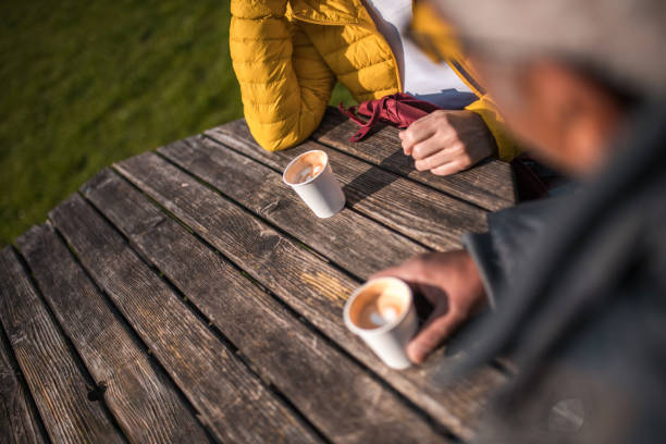 Elderly male and female in casual attire sitting at a wooden table, enjoying hot beverages outdoors, reflecting an active lifestyle. Focus on hands and cups.