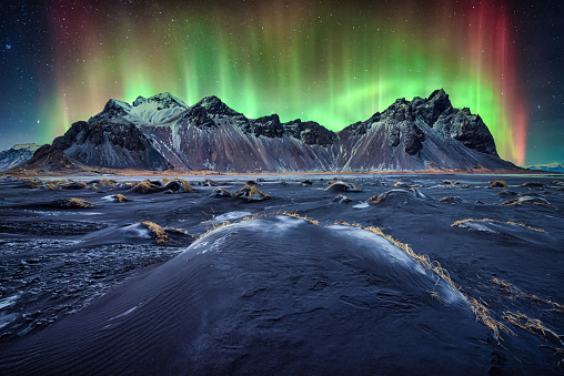 Green and red northern lights glowing over Vestrahorn mountain at night.  Great wind rippled black sand beach. Location famous place Stokksnes cape, Vestrahorn , Iceland, Europe