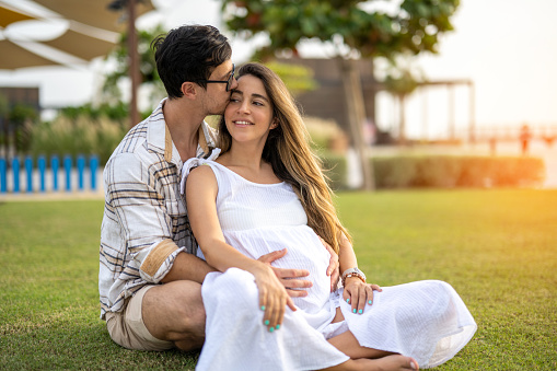 Beautiful pregnant woman sitting in her husband's arms on the grass in the park. Handsome man kissing his wife