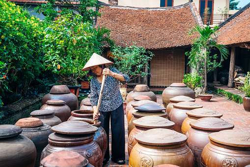 A Vietnamese woman in traditional conical hat stands among large clay pots in a serene courtyard near Hanoi, illustrating cultural heritage.