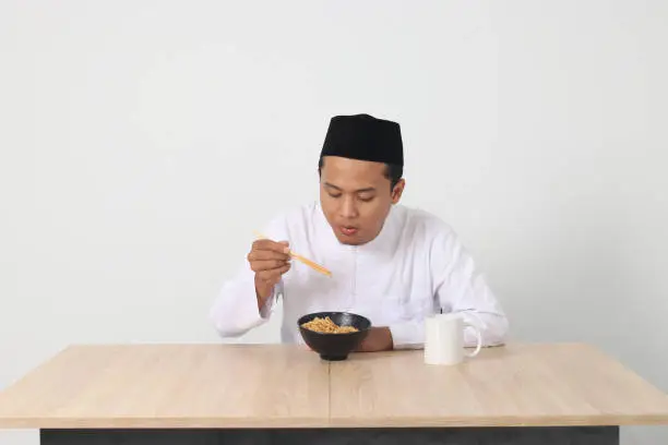 Portrait of attractive Asian muslim man eating tasteful instant noodles with chopsticks served on bowl. Iftar and pre dawn meal concept. Isolated image on white background