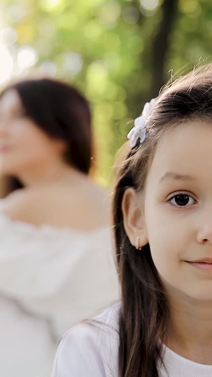 Happy face of a child in the rays of the sun. Portrait of a girl in the foreground and her mother in the blurred background in a park. Mom and daughter. Vertical video