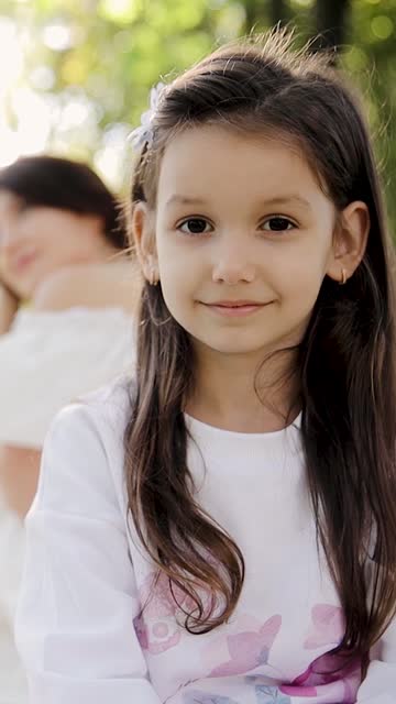 Happy cute affectionate adopted little child girl in foreground and foster mother in blurred background in park. Foster home adoption concept, steadicam shot. Vertical video