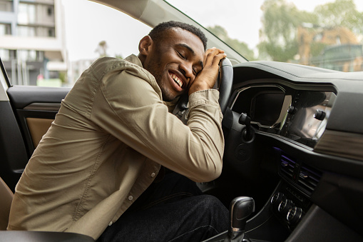 Delighted african american man sitting in the driver's seat, hugging the steering wheel of his new car, closing his eyes and smiling.