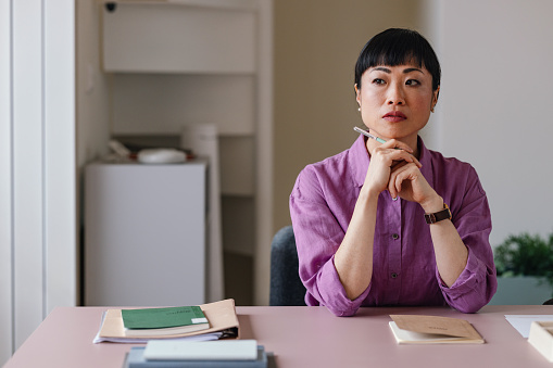 A pensive Asian businesswoman sitting at her desk with a thoughtful expression.