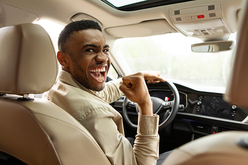 Joyful african american man holding steering wheel shaking fist while sitting in car at car dealership.Own car. High quality photo