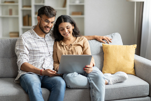 Loving happy indian couple sitting on couch at home, embracing, using laptop computer, young man and woman watching movie online, copy space