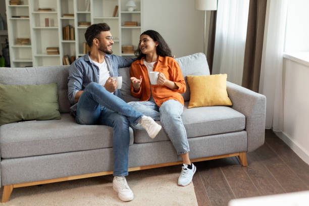 istock Happy indian couple relaxing and drinking coffee, sitting on couch at home, talking while resting with hot drinks in cups at modern living room, free space 2092374719