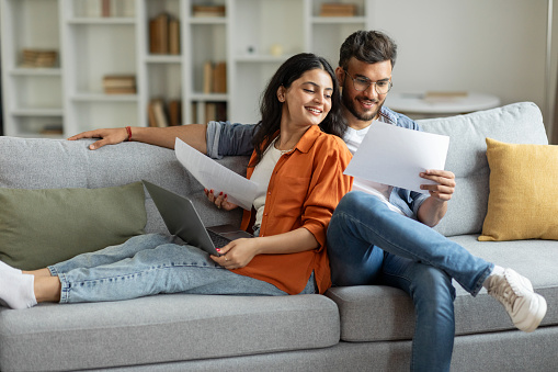 Happy indian couple checking documents, spouses reading insurance agreement or property certificate, husband and wife sitting on couch in living room, copy space