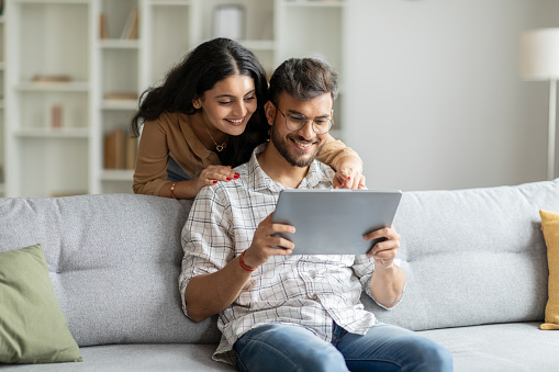 Happy indian man and woman sitting on couch, hugging and using digital tablet, looking at pad screen and smiling, using newest gadget at home interior