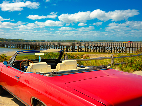 Idyllic Retro-style Photo of Summer Beach Seascape with a convertible red antique car parked under the bridge on a bright sunny day with cloudy sky