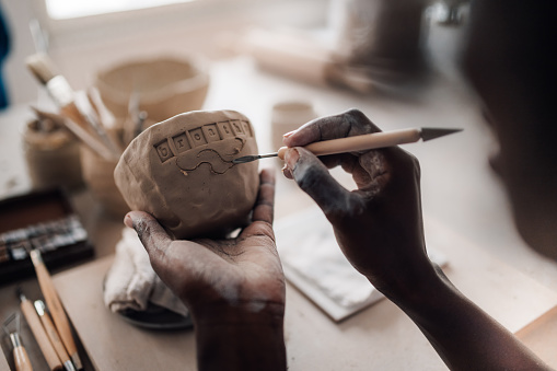 Close up of a multicultural craftswoman's hands decorating pottery and clay work at pottery class in modern studio. Cropped picture of an african american pottery course student decorating earthenware