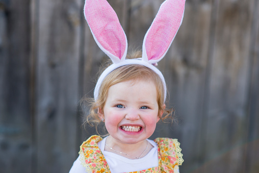 Cute little girl is smiling a toothy smile outdoors in front of a barn while wearing a yellow dress and costume rabbit ears for Easter celebrations with her family in the Spring.