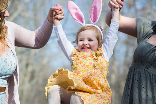 Cute little girl in a yellow Easter dress is laughing and smiling as she is being lifted up by her mother and aunt while on a morning Easter walk. She is wearing costume rabbit ears like the Easter bunny.