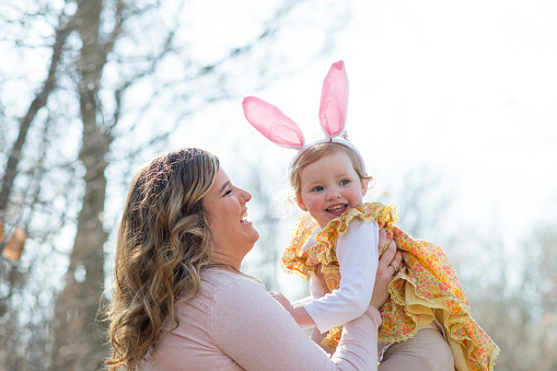 Beautiful mother is holding up her young daughter playfully outdoors. The little girl is wearing a yellow Easter dress and costume rabbit ears like the Easter bunny for the holiday.