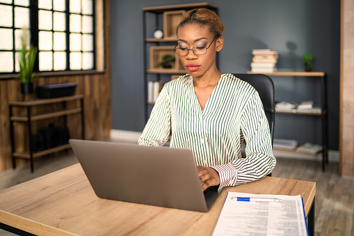 A serious African-American woman is sitting at her desk typing on her laptop, dressed in a smart business shirt and wearing eyeglasses she is concentrating on her work in a modern office