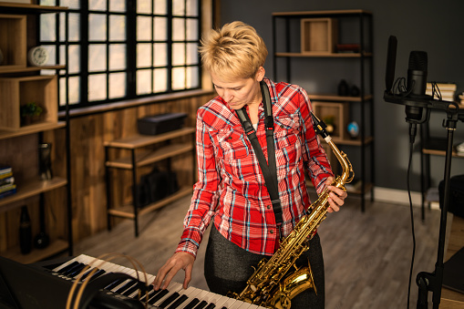 A talented female saxophonist is holding her saxophone while also playing on a keyboard in a music sound recording studio