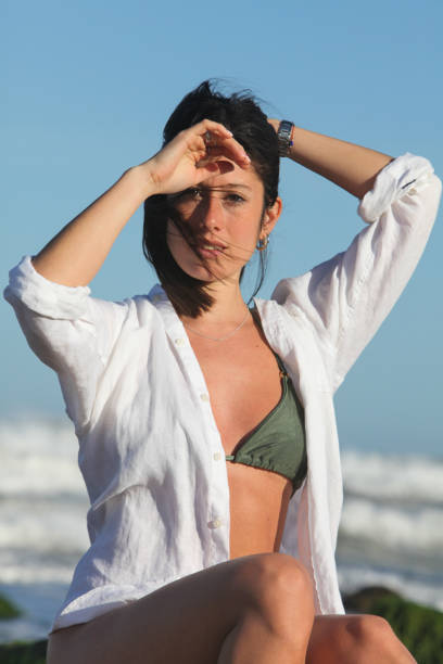 pretty young woman in bikini and white shirt  sitting by the ocean