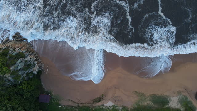 Slowmo aerial top-down view of a sandy beach being hit by foamy storm waves