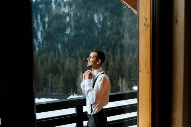 istock Young man wearing suspenders, buttoning his magpie while standing on a balcony with a view of snow-capped mountains. 2092624085