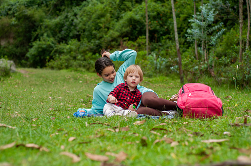 A boy and a girl are seated on a grassy field, surrounded by the freshness of springtime greenery. The girl is attentively helping the younger boy with a task, while a bright pink backpack rests beside them, suggesting a day of adventure and exploration in the great outdoors.