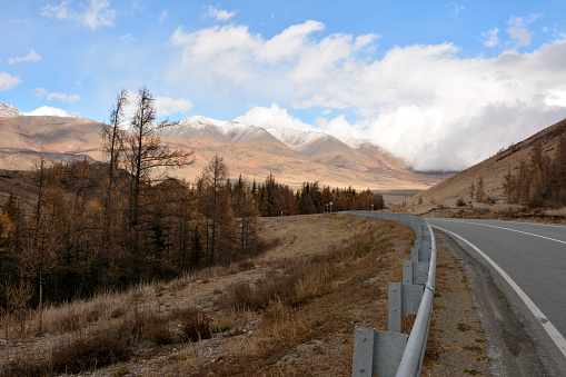 An asphalt road with metal fences descends from the mountain into a picturesque autumn valley at the foot of snow-capped mountains on a sunny autumn day. Chuisky tract, Altai, Siberia, Russia.