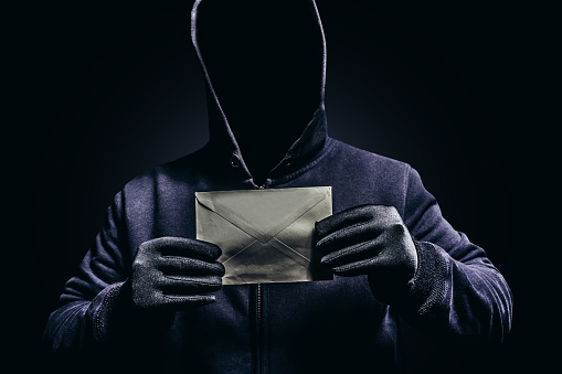 Photo of scary shaded hooded man holding paper letter on dark background.