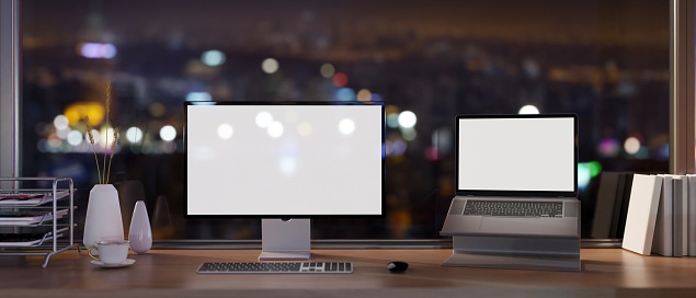 A modern skyscraper office at night with a computer and white-screen laptop mockup on a desk against the glass window with a night city view. 3d render, 3d illustration