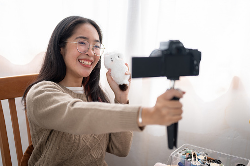 A smiling, cheerful young Asian female content creator is talking with the camera, showing her handmade plushie at her sewing table at home.