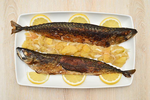 Roasted fish and potato salad with onion and lemon slices for decoration on the brown wooden table. Top view.