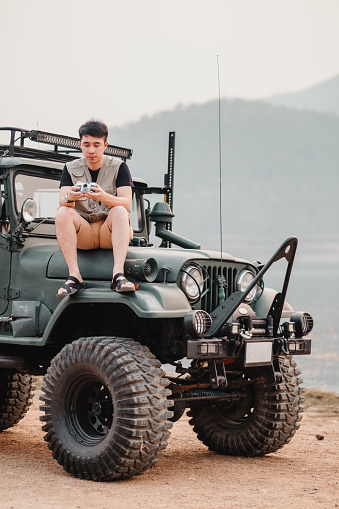 Young man sits on a heavily modified car, holding a camera, ready for an off-road photography adventure.