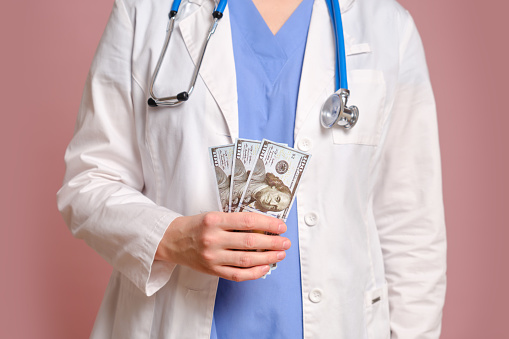 Woman doctor holding money in us dollars in her hands, studio pink background. Nurse in uniform with stethoscope on red studio background