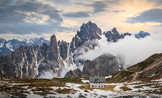 Panoramic view of rugged mountain range in Dolomites, Italy, with  snow, mountain hut and footpath at sunset;  sky in background