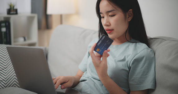Selective focus, Young asian woman sitting on sofa holding credit card making online payment for purchase in web store using laptop