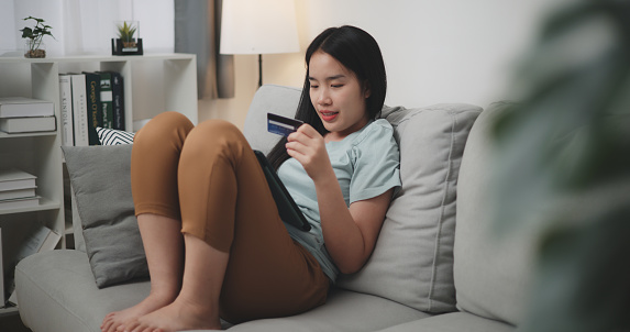 Selective focus, Happy young woman sitting on sofa using credit card with mobile phone for online shopping cashless in living room at home