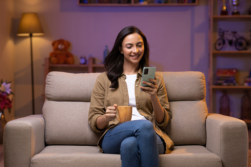 Young girl drinking coffee while using phone sitting on sofa at home