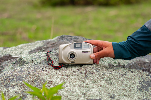 A close-up of a hand placing an old analog camera on a moss-covered rock, surrounded by nature