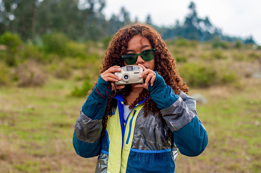 A young woman in sunglasses and sportswear takes a photo with an old camera, standing in a field