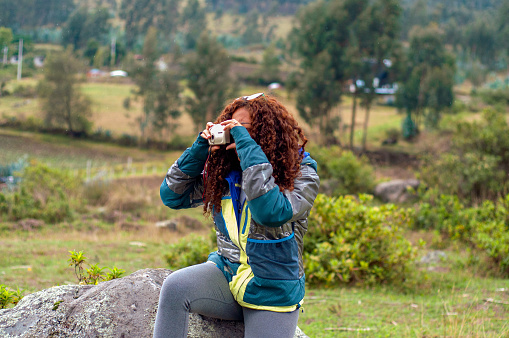 A woman sits on a rock, focusing her camera on a subject, surrounded by greenery