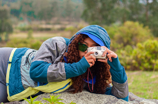 A young woman lies on her stomach outdoors, taking a picture with a small camera, wearing a hooded jacket and sunglasses. Her curly hair cascades beside her face, implying a relaxed yet focused moment as she engages with her surroundings. The cloudy weather creates a soft light, ideal for outdoor photography.