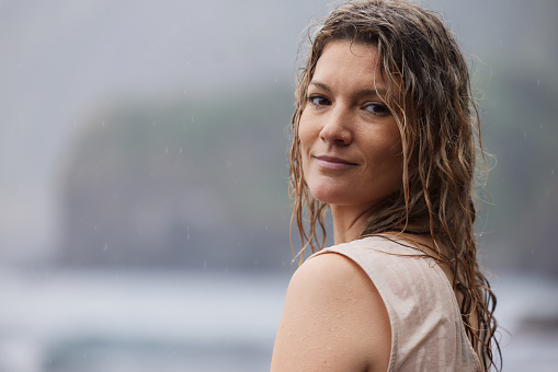 Portrait of carefree woman with wet hair standing on the beach during rainy day and looking at camera. Copy space.